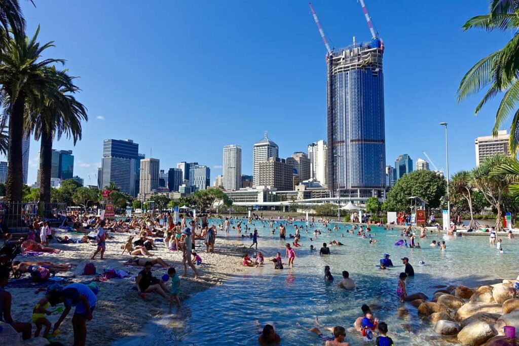 Billede af Street Beach i Brisbane på en solrig dag, med mange mennesker, der nyder stranden og vandet, og byens skyline i baggrunden.