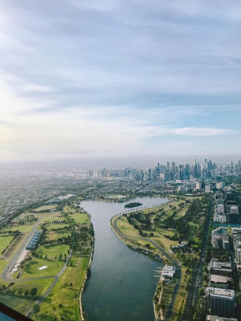 Luftfoto af en park i Melbourne med en stor sø i midten, omgivet af grønne områder, sportsbaner og veje, med byens skyline i baggrunden.