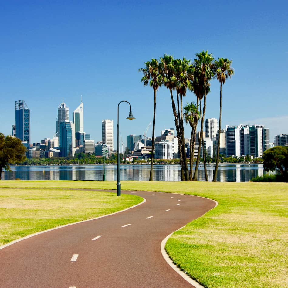 Perth, Western Australia, med en snoet gang- og cykelsti i forgrunden, der fører gennem en park med palmetræer, og byens skyline i baggrunden ved bredden af en flod.