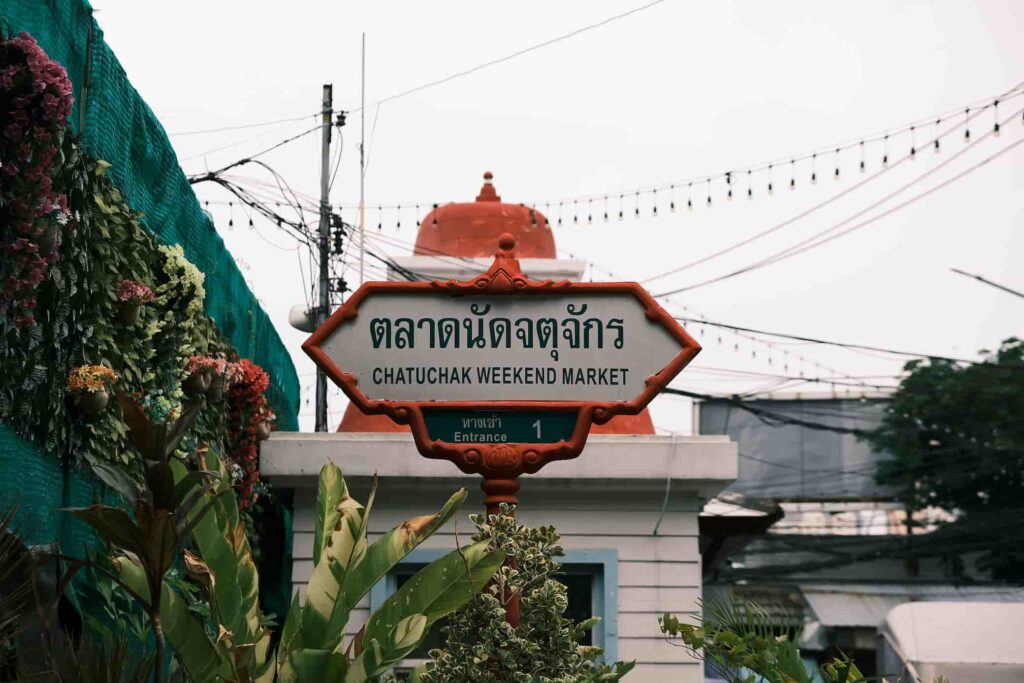 Chatuchak Weekend Market i Bangkok, Thailand, viser den velkendte markedslogo med levende farver og thailandsk skrift, omgivet af livlige boder og travle besøgende.